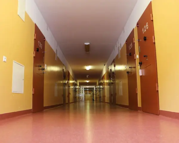 Długi korytarz z rzędem drzwi, typowy dla zakładu karnego dla kobiet. Jasne ściany i drzwi z numerami cel.