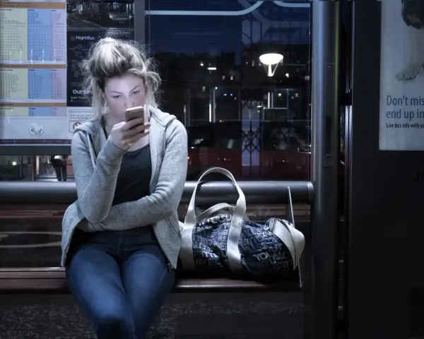 Dziewczyna na przystanku nocnego autobusu, pochłonięta telefonem, ignoruje uporczywe niepokojenie. Obok torba.