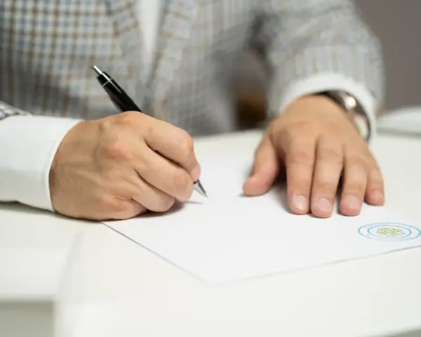 Mężczyzna w eleganckim garniturze podpisuje ważny dokument. Jego ręka pewnie trzyma długopis, a na papierze widnieje logo firmy.