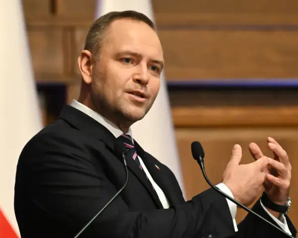 Mężczyzna w garniturze przemawia, gestykulując rękami. W tle biało-czerwona flaga, symbol prawa i sprawiedliwości.