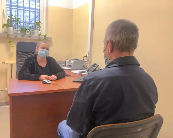 Psycholog więzienny w masce rozmawia z osadzonym. W tle okno z kratami i rośliny.