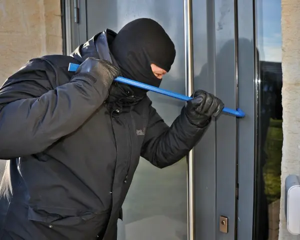 Osoba w kominiarce i rękawiczkach próbuje wyważyć drzwi łomem. To przykład kradzieży z włamaniem.