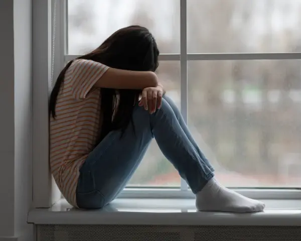 Dziewczyna siedzi skulona przy oknie, pogrążona w smutku. Obraz symbolizuje traumę po przestępstwach na tle seksualnym.