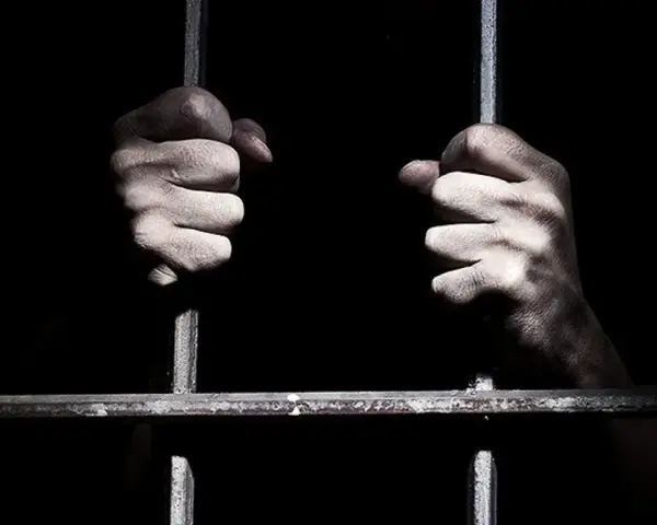 Dłonie zaciskają się na zimnych prętach krat. Czy w więzieniu jest ciężko? Ciemność i stal sugerują odpowiedź.