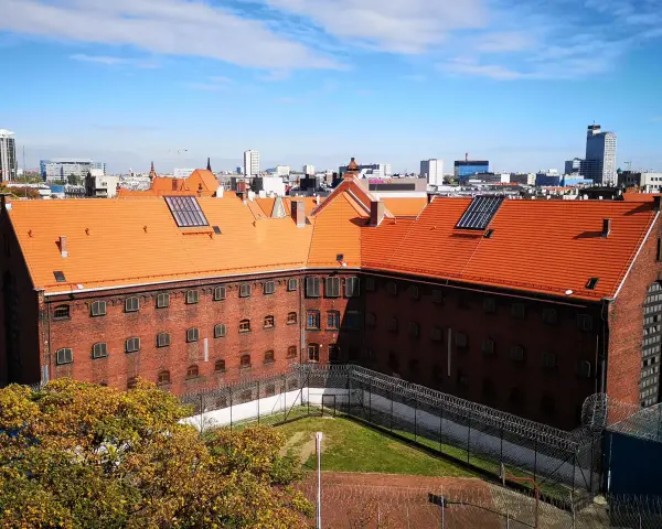 Widok na ceglany budynek z pomarańczowym dachem, otoczony ogrodzeniem z drutu kolczastego. W tle miejski krajobraz z wieżowcami. To zakład karny Służewiec.