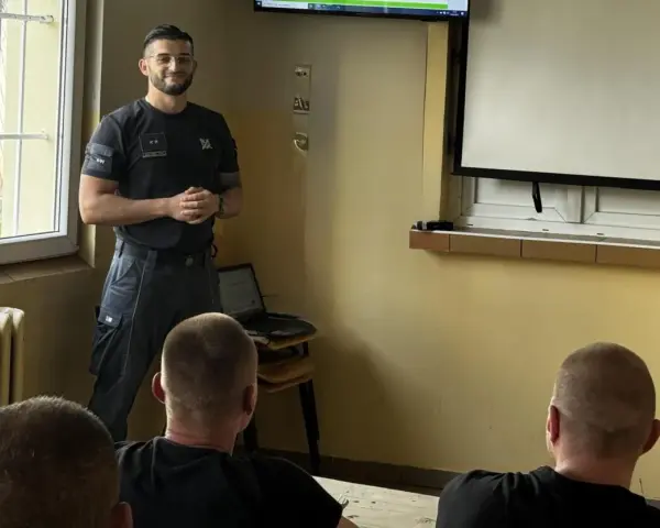Strażnik więzienny prowadzi szkolenie w sali przypominającej izolatkę. Na ekranie prezentacja, a przed nim słuchacze.
