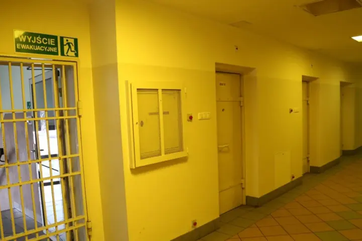 Korytarz więzienny z kratami i drzwiami cel. Nad drzwiami znak "Wyjście ewakuacyjne".