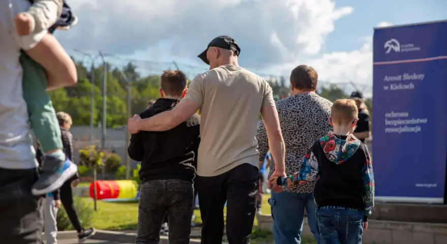 Rodzina idzie w stronę aresztu. Dzieci trzymają się za ręce, a dorośli obejmują się nawzajem. W tle widać ogrodzenie i tablicę informacyjną. To obraz matki za kratami, która spotyka się z bliskimi.