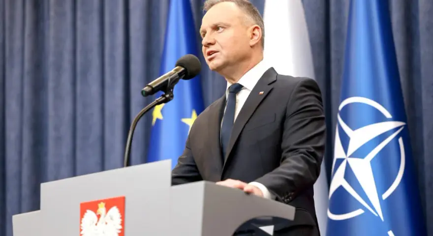 Prezydent Andrzej Duda przemawia przy mównicy z godłem Polski. Za nim flagi UE i NATO. Może to być związane z ułaskawieniem prezydenta.