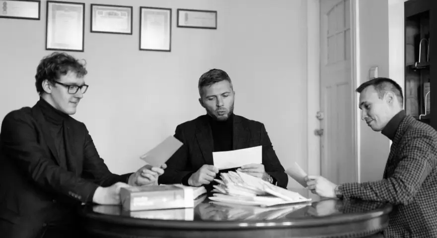 Trzech mężczyzn przy okrągłym stole analizuje dokumenty. Jeden z nich, z okularami, podaje kopertę. W środku siedzi mężczyzna z zarostem, przeglądający stos papierów, a po jego prawej stronie siedzi mężczyzna w kratkę, również z dokumentem. W tle wiszą...