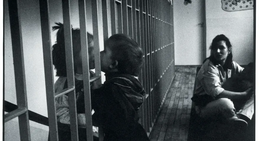Dzieci przy kratach, zastanawiamy się, czy zabierać dziecko do zakładu karnego. Kobieta siedzi w tle.
