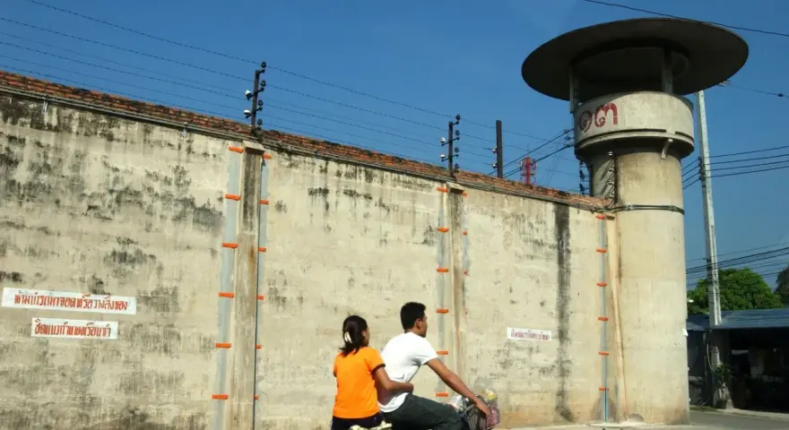 Para rowerzystów mija wysoki mur z drutem kolczastym, przypominający więzienie w Tajlandii. Obok stoi wieża strażnicza.
