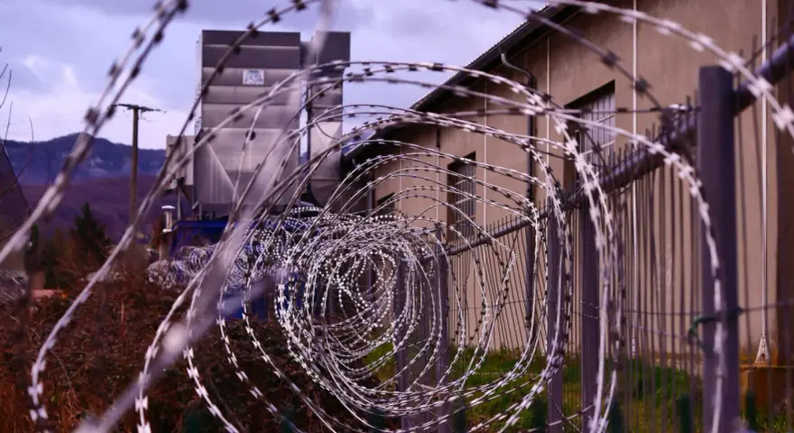 Kolczasty drut tworzy spiralę przed budynkiem. W tle widać góry i przemysłową konstrukcję. Może to miejsce, gdzie zapadały decyzje komisji penitencjarnej.