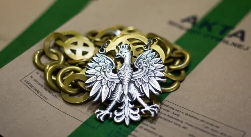 Srebrny orzeł na złotym łańcuchu, leżący na aktach. Symbolizuje prekluzja dowodowa w polskim prawie.
