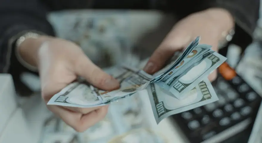 Dłonie liczą banknoty 100-dolarowe. Obok leży kalkulator, sugerując obliczanie kwoty, być może jako kara pieniężna za wyrządzoną krzywdę.