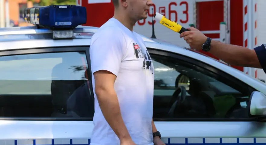 Policjant przeprowadza procedurę badania alkomatem. Mężczyzna dmucha w urządzenie przy radiowozie.