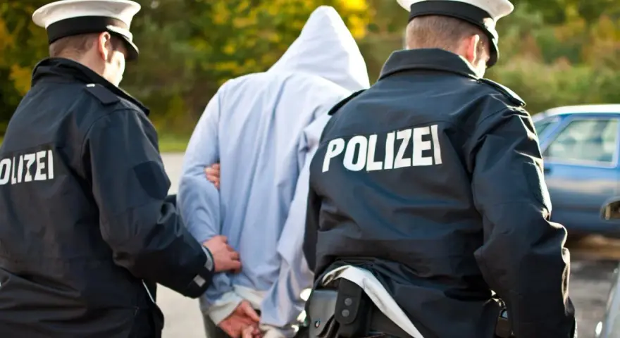 Dwóch policjantów w mundurach z napisem "POLIZEI" prowadzi zatrzymanego mężczyznę w kapturze. Zastanawiasz się, ile trwa areszt w Niemczech?