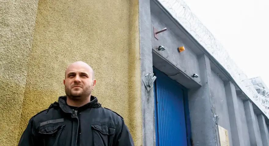 Łysy mężczyzna w czarnej kurtce stoi przed budynkiem z niebieskimi drzwiami i drutem kolczastym. Może to być miejsce, gdzie słychać gwara przestępcza.