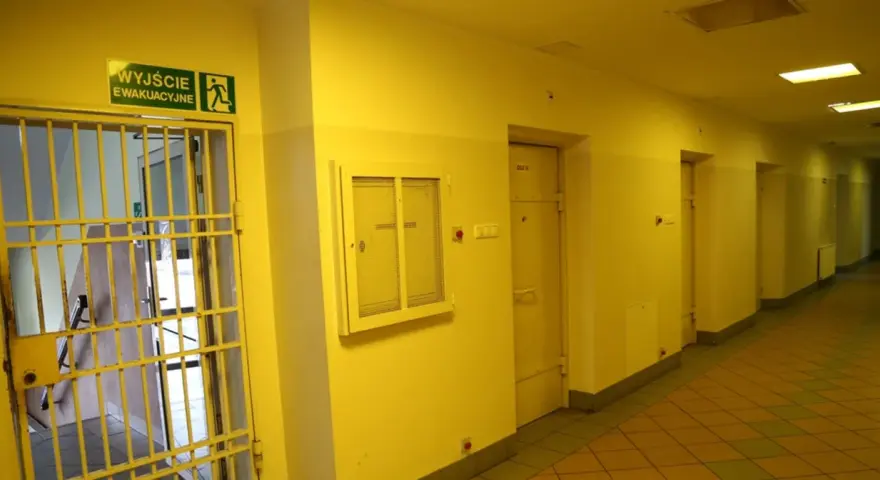 Korytarz więzienny z kratami i drzwiami cel. Nad drzwiami znak "Wyjście ewakuacyjne".