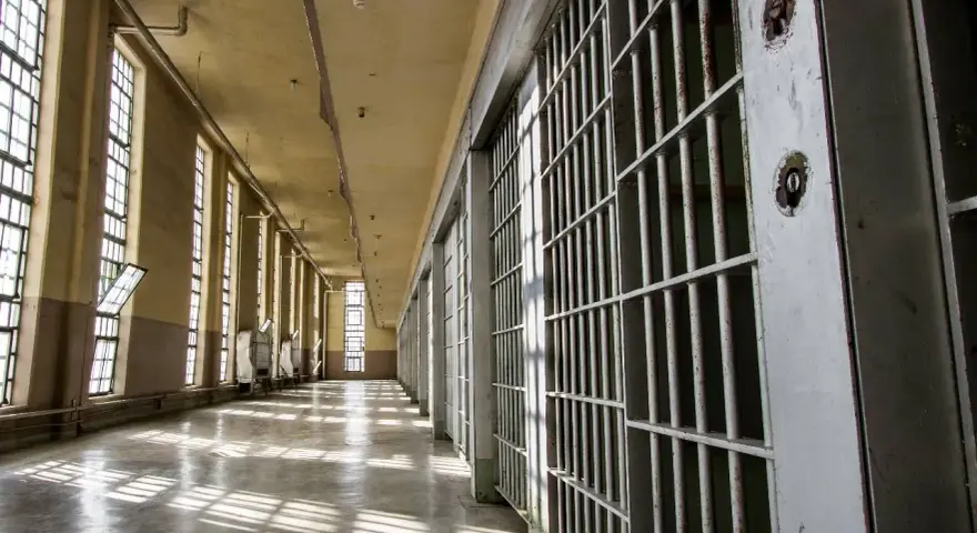 Korytarz więzienny z celami po prawej i oknami po lewej. Światło wpada przez okna, tworząc cienie na podłodze. Zastanawiasz się, po jakim czasie więzień może dzwonić?
