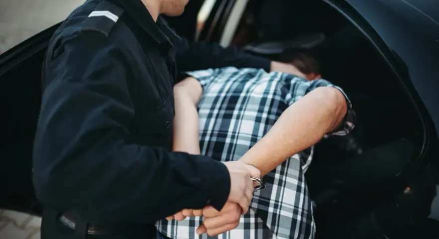 Policjant zakłada kajdanki zatrzymanemu mężczyźnie w kratkę, który nie stawia oporu. To jego dobrowolne poddanie się karze.
