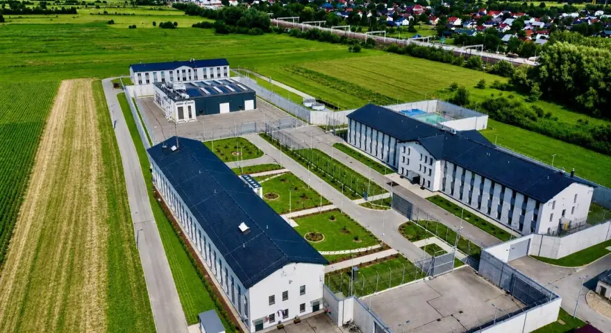 Nowoczesny kompleks budynków otoczony ogrodzeniem, przypominający najnowsze więzienie w Polsce, otoczony polami i domami mieszkalnymi.