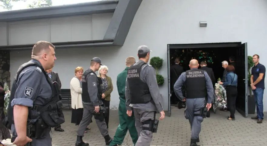 Strażnicy więzienni eskortują więźnia na pogrzeb. Widać ich w mundurach przed budynkiem, gdzie odbywa się ceremonia.