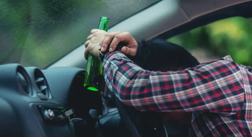 Mężczyzna w kratę, z butelką w ręku, opiera się o kierownicę. Pytanie: po ilu latach występuje przedawnienie za jazdę pod wpływem alkoholu?