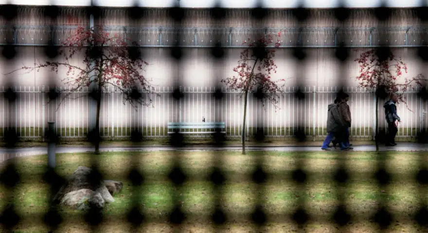 Więzienie w Niemczech. Ludzie spacerują po dziedzińcu, drzewa z czerwonymi liśćmi, ławka, kamień i ogrodzenie.