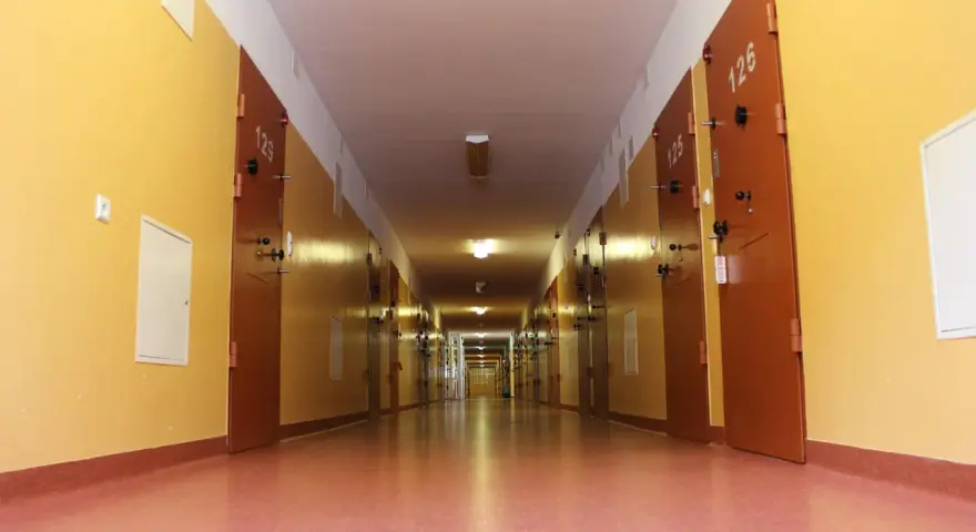 Długi korytarz z rzędem drzwi, typowy dla zakładu karnego dla kobiet. Jasne ściany i drzwi z numerami cel.