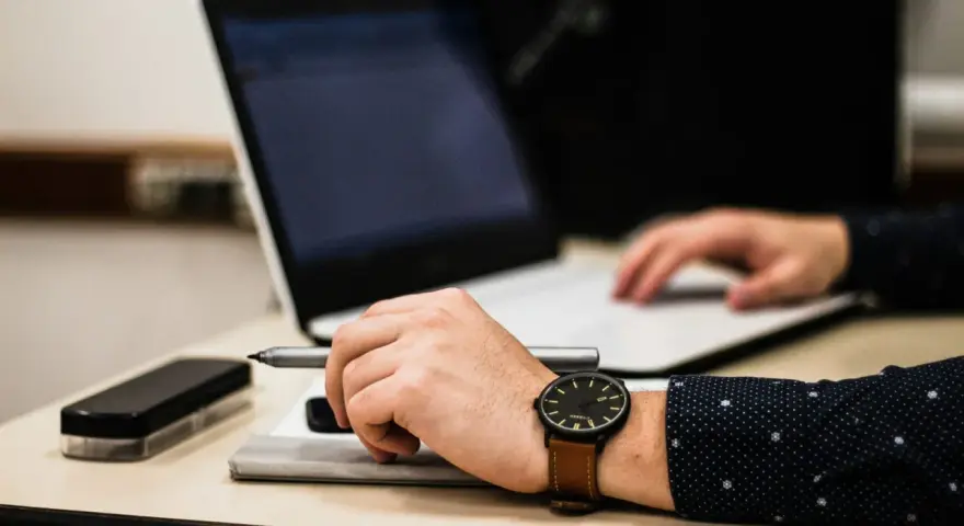 Dłoń z zegarkiem trzyma długopis nad dokumentem, obok laptop. Czas na skargę na przewlekłość postępowania opłatę.