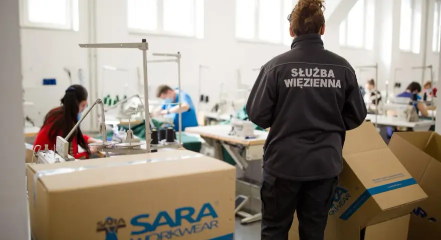 Pracownicy wykonują pracę w więzieniu, szyjąc odzież. Nadzorca w kurtce z napisem "Służba Więzienna" obserwuje proces.