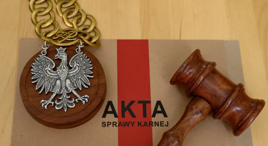 Na brązowej teczce z napisem "AKTA SPRAWY KARNEJ" leży drewniany młotek sędziowski i medalion z orłem RP.