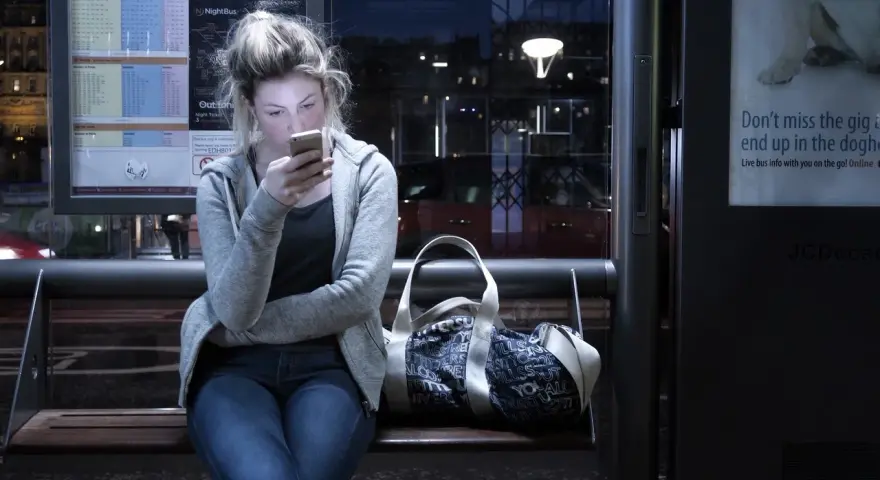 Dziewczyna na przystanku nocnego autobusu, pochłonięta telefonem, ignoruje uporczywe niepokojenie. Obok torba.