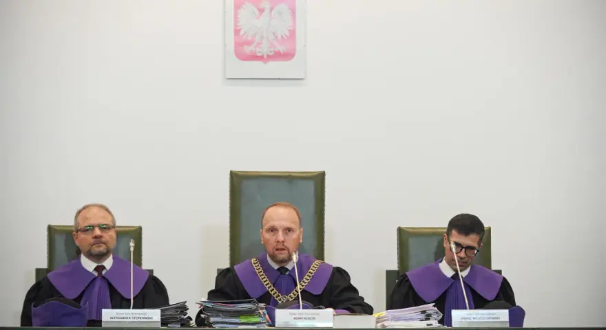 Trzech sędziów w togach podczas jawnego posiedzenia sądu. Na ścianie wisi godło Polski.