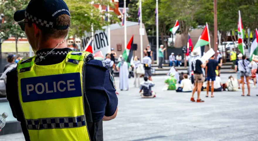 Policjant w kamizelce odblaskowej obserwuje tłum z flagami. Sytuacja przypomina zatrzymanie nieletniego przez policję, choć nie widać konkretnego zdarzenia.