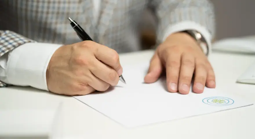 Mężczyzna w eleganckim garniturze podpisuje ważny dokument. Jego ręka pewnie trzyma długopis, a na papierze widnieje logo firmy.