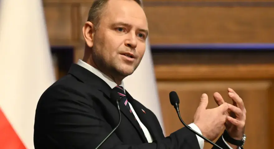 Mężczyzna w garniturze przemawia, gestykulując rękami. W tle biało-czerwona flaga, symbol prawa i sprawiedliwości.
