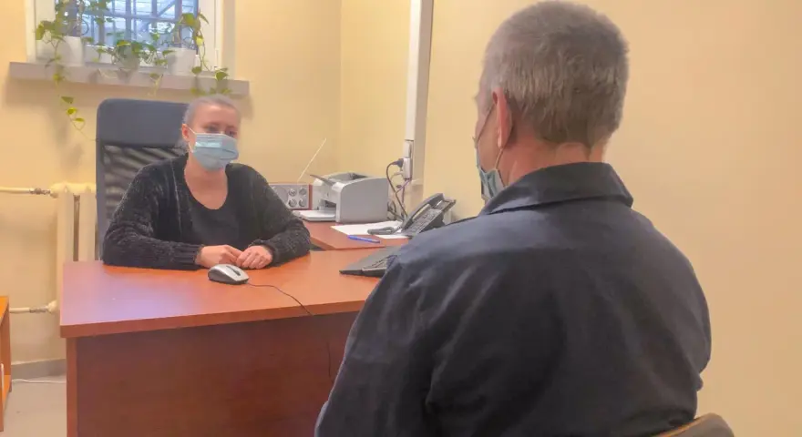 Psycholog więzienny w masce rozmawia z osadzonym. W tle okno z kratami i rośliny.