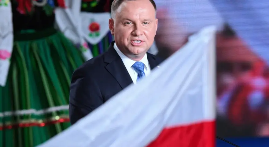 Prezydent Andrzej Duda w garniturze i krawacie, na tle polskiej flagi i tradycyjnego stroju. Ile trwa ułaskawienie prezydenta?