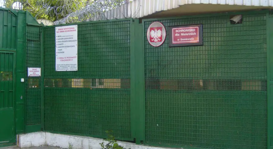 Zielona brama z drutem kolczastym i znakiem "Schronisko dla Nieletnich w Dominowie". To miejsce, gdzie młodzież potrzebuje wsparcia, a nie kary.