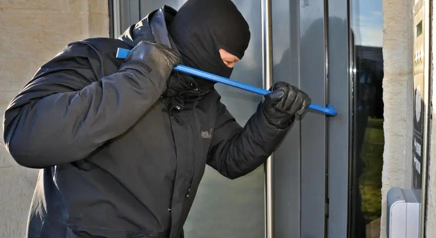Osoba w kominiarce i rękawiczkach próbuje wyważyć drzwi łomem. To przykład kradzieży z włamaniem.