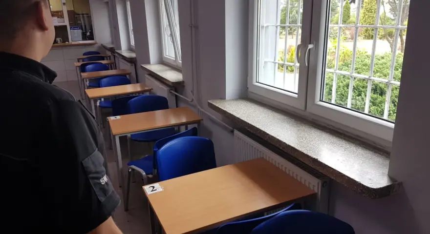 Rzędy pustych ławek i krzeseł w sali lekcyjnej. Za kratami okien widać zieleń. To może być sala egzaminacyjna w więzieniu na Białołęce.