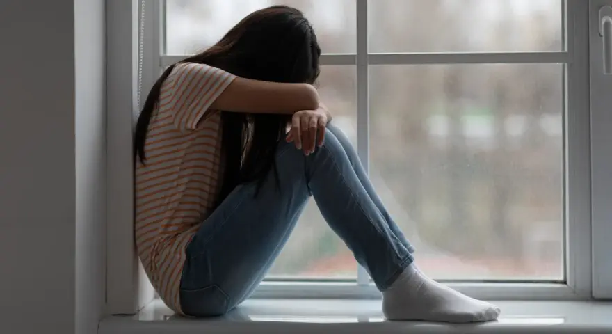 Dziewczyna siedzi skulona przy oknie, pogrążona w smutku. Obraz symbolizuje traumę po przestępstwach na tle seksualnym.
