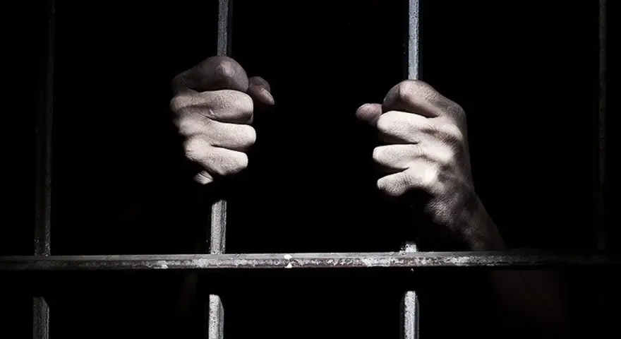 Dłonie zaciskają się na zimnych prętach krat. Czy w więzieniu jest ciężko? Ciemność i stal sugerują odpowiedź.