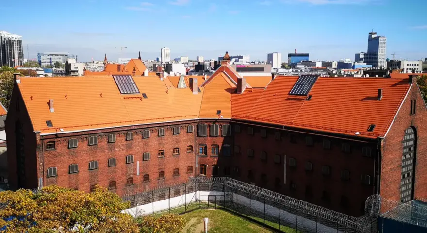 Widok na ceglany budynek z pomarańczowym dachem, otoczony ogrodzeniem z drutu kolczastego. W tle miejski krajobraz z wieżowcami. To zakład karny Służewiec.