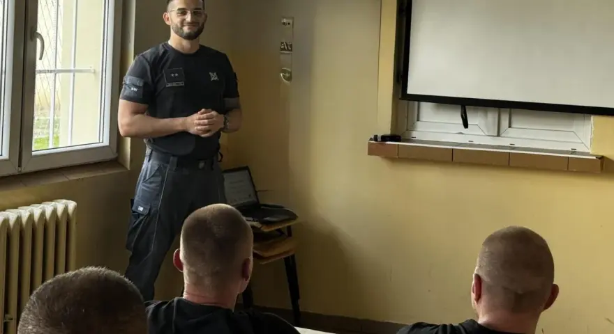 Strażnik więzienny prowadzi szkolenie w sali przypominającej izolatkę. Na ekranie prezentacja, a przed nim słuchacze.