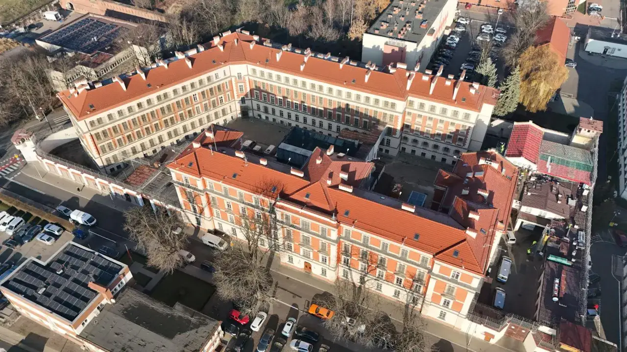 Widok z lotu ptaka na zakład karny Służewiec. Budynki z czerwonymi dachami i licznymi oknami otoczone murem z drutem kolczastym.