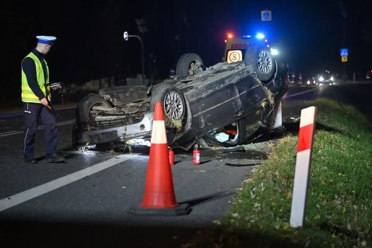 Policjant dokumentuje wypadek. Przewrócony samochód na drodze, w tle karetka. Może to być podstawa do art. 177 k.k. i kara grzywny.
