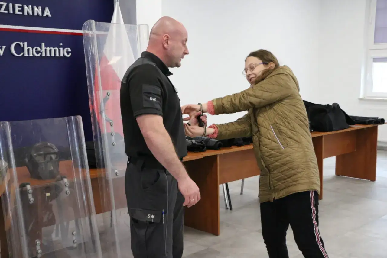 Pierwsze widzenie w zakładzie karnym. Kobieta zakłada kajdanki mężczyźnie. W tle tarcze ochronne i hełm.